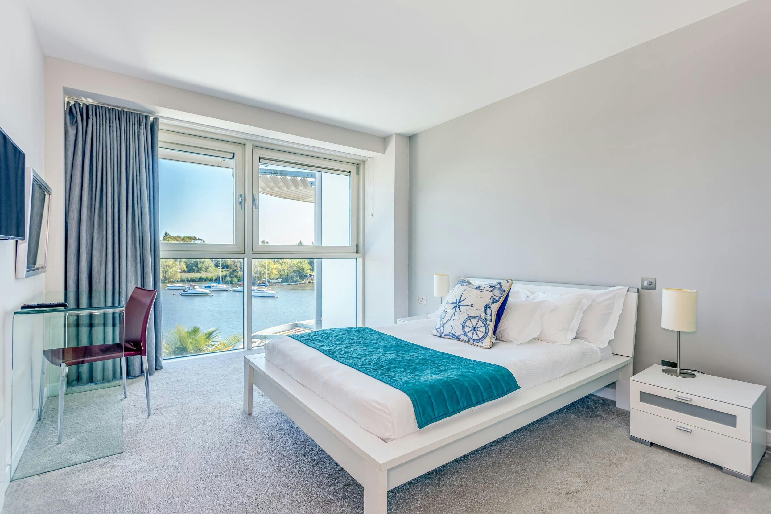 A bright bedroom with a white bedframe and full-height glass onto the river