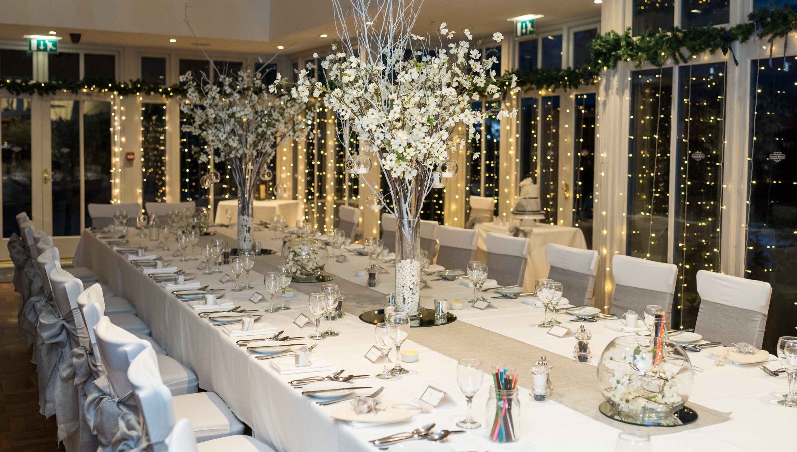 A long banqueting table under fairy lights with tall blossom trees down its length