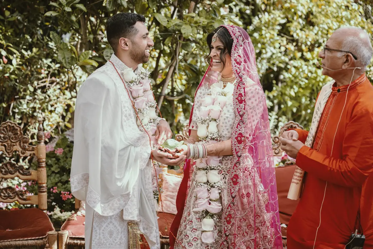 A Hindu ceremony under garden trees, groom and bride laughing mid-exchange