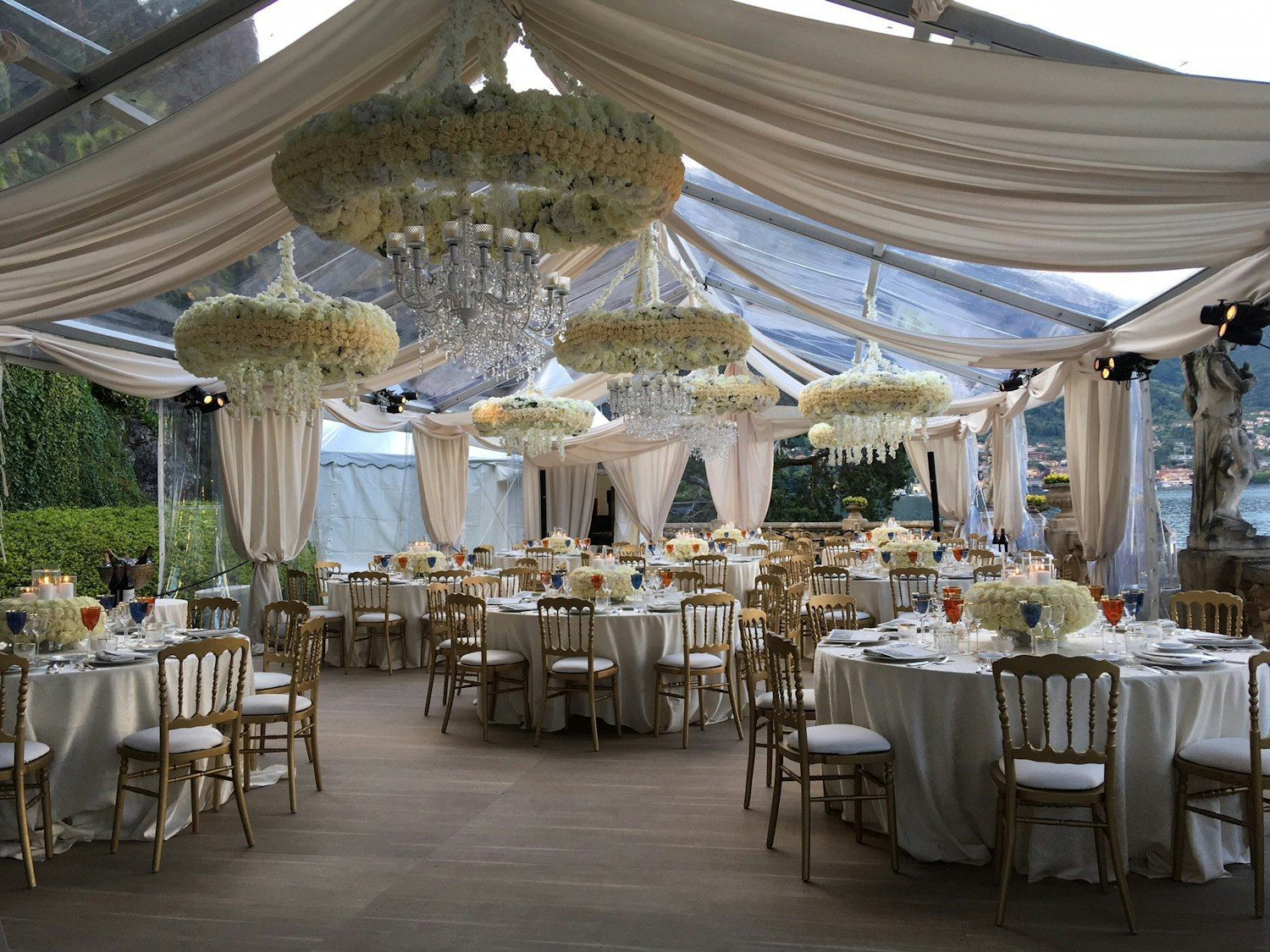 A wedding reception under a clear marquee on the lake terrace, floral chandeliers and gold chairs, Villa del Balbianello