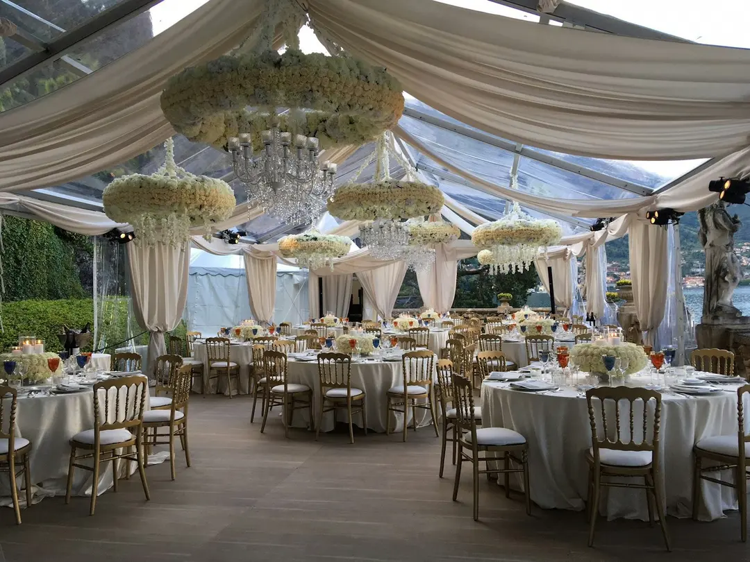 A wedding reception under a clear marquee on the lake terrace, floral chandeliers and gold chairs, Villa del Balbianello