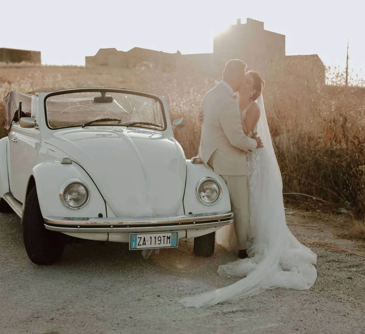 A bride and groom kissing beside a vintage white Volkswagen Beetle convertible at golden hour