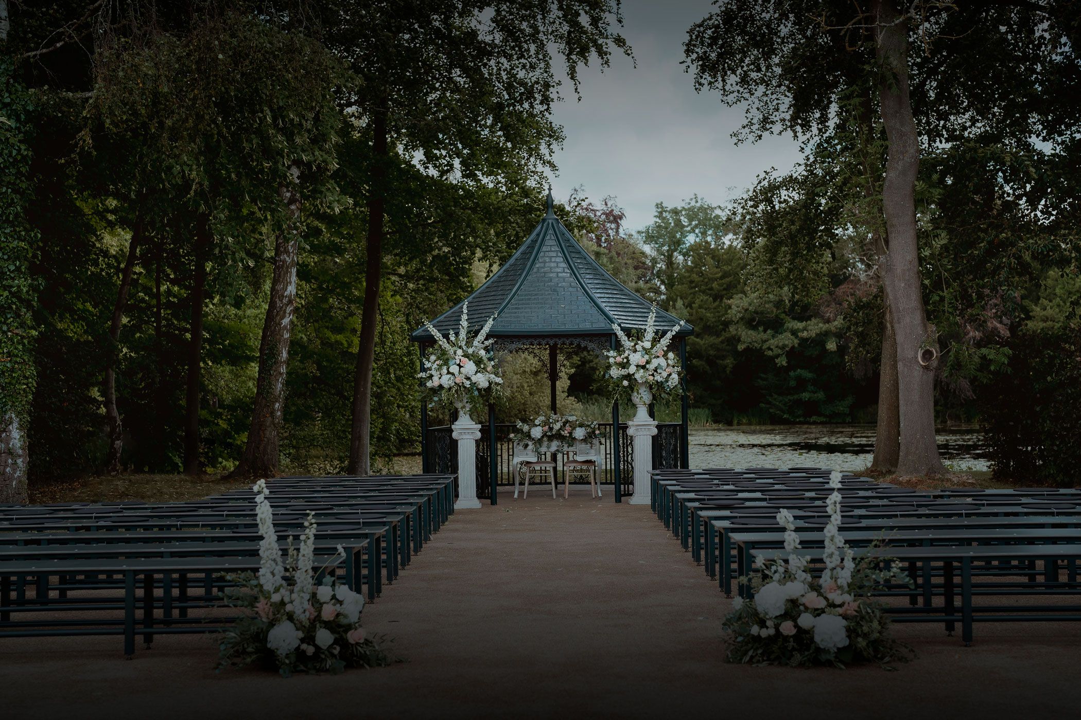An outdoor ceremony set beneath the trees, an aisle of benches leading to a floral-dressed pavilion