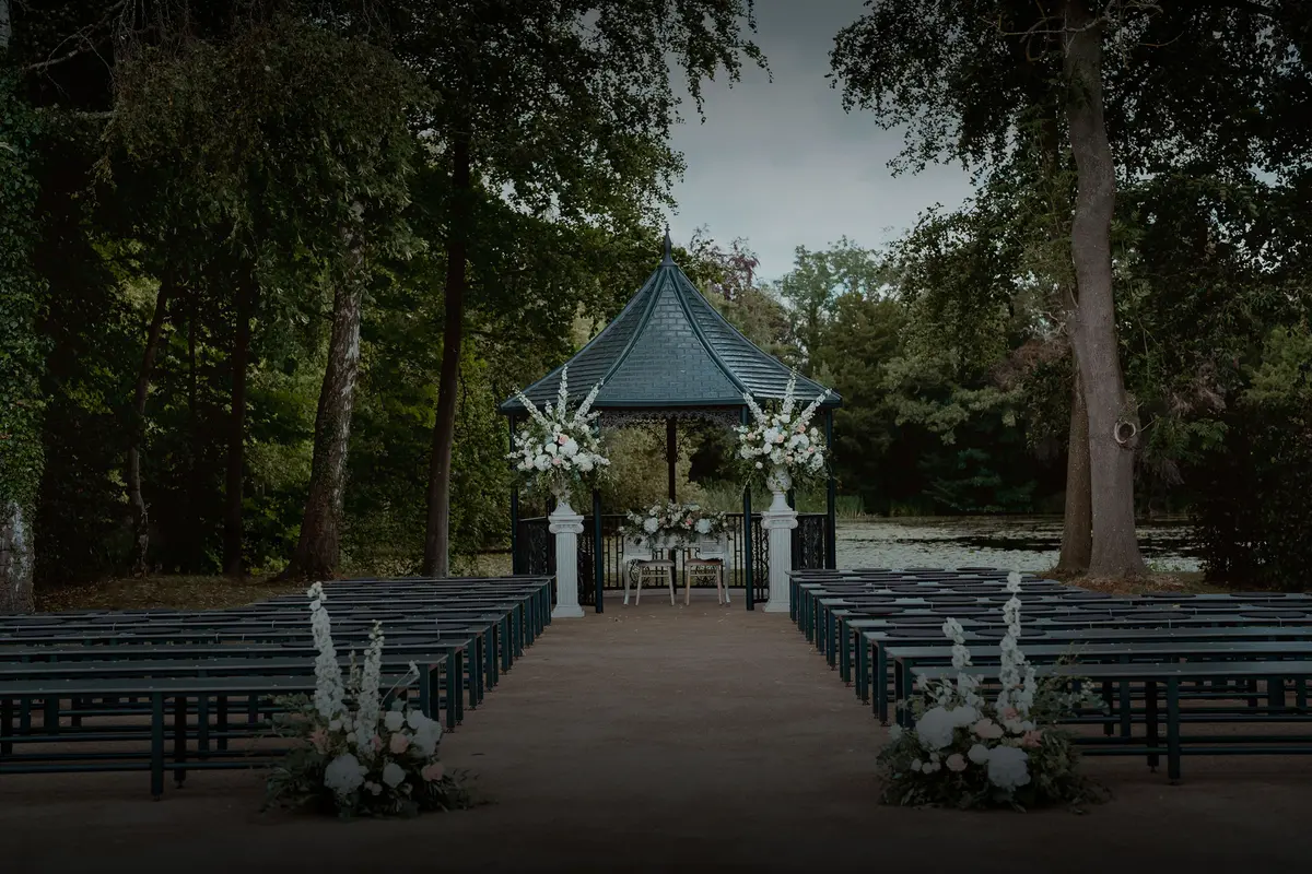 An outdoor ceremony set beneath the trees, an aisle of benches leading to a floral-dressed pavilion, Hengrave Hall, Bury St Edmunds, England