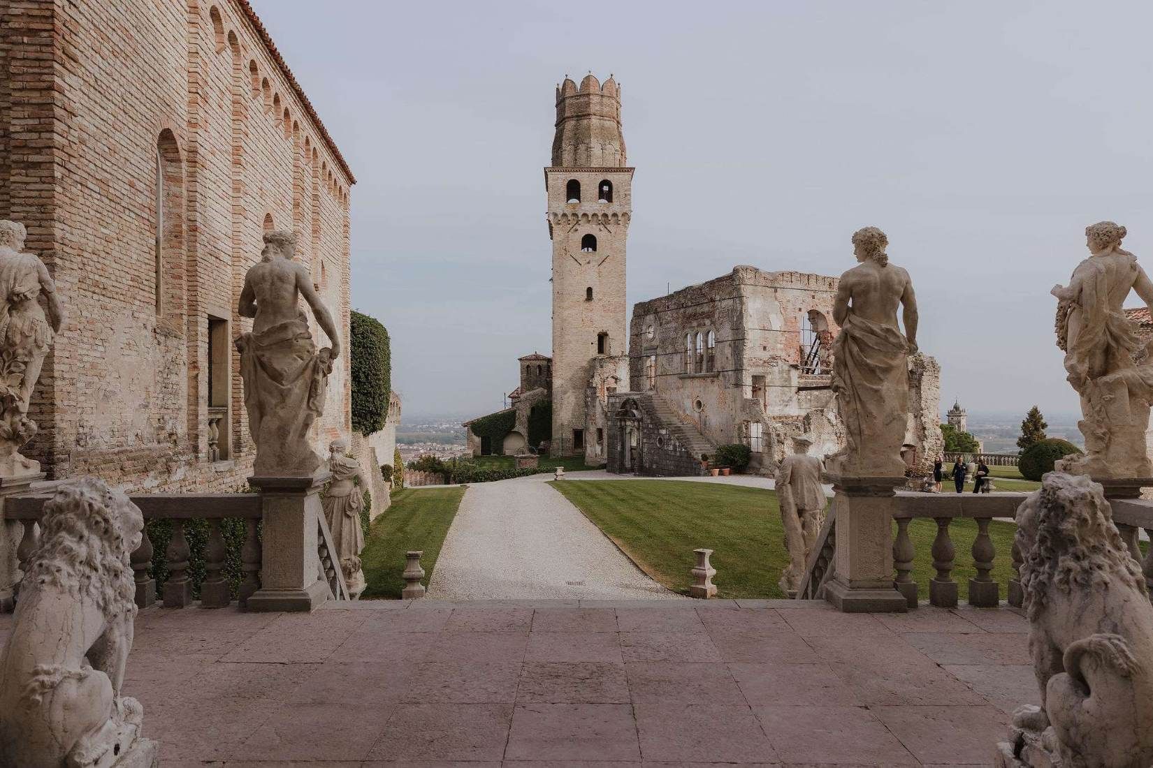 A ruined medieval tower and cloister on a Veneto hilltop, guarded by weathered stone lion statues