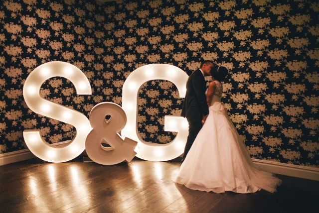 A bride and groom beside a lit-up "S & G" sign at their reception