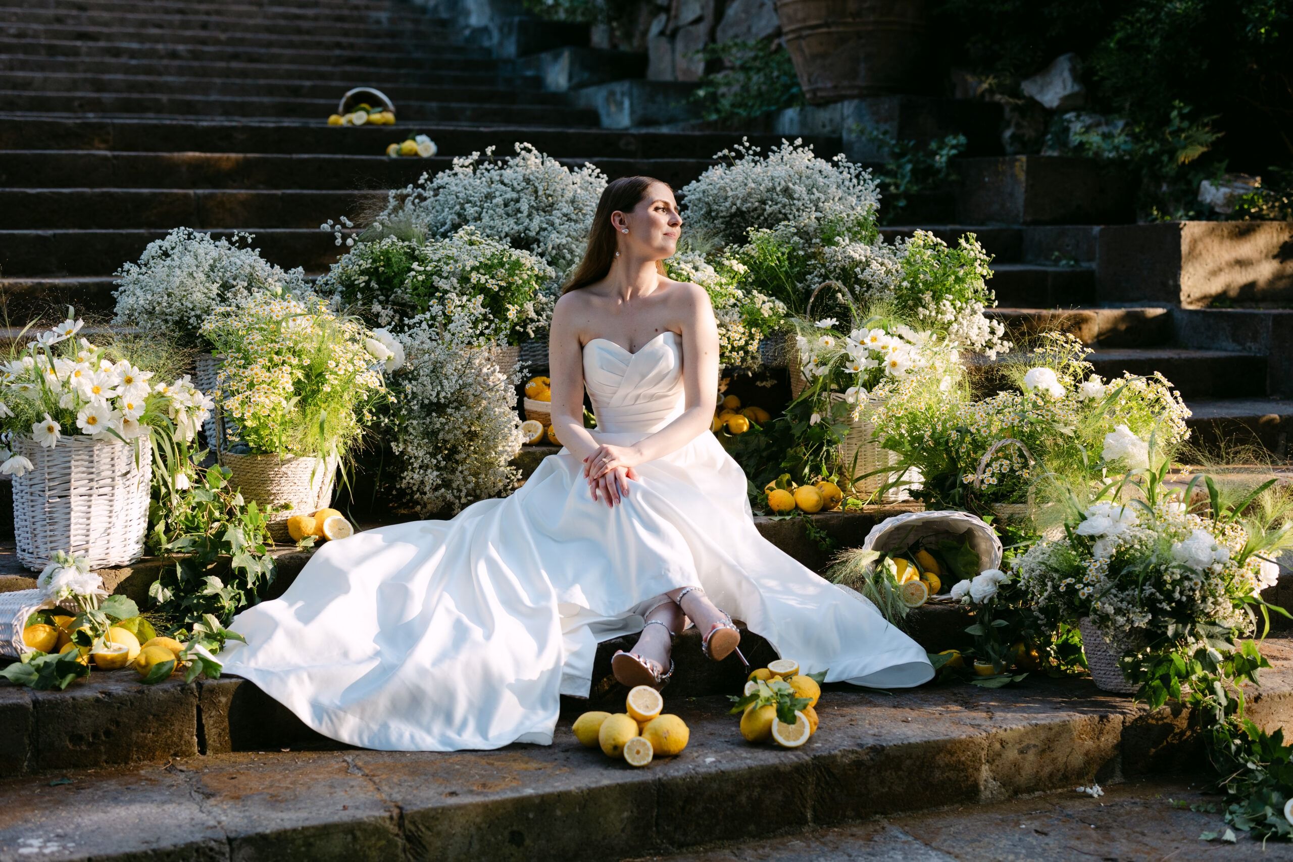 The bride alone on stone steps banked with baskets of lemons