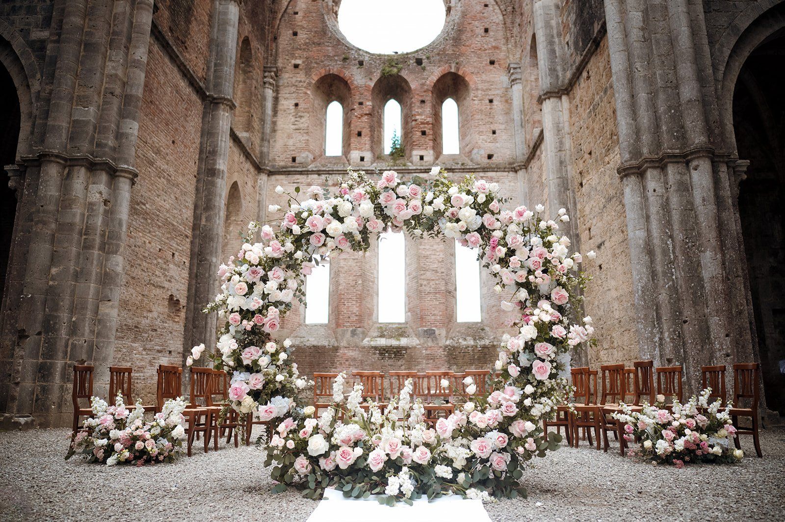 A circular arch of pink and white roses inside the roofless nave of San Galgano Abbey, Siena