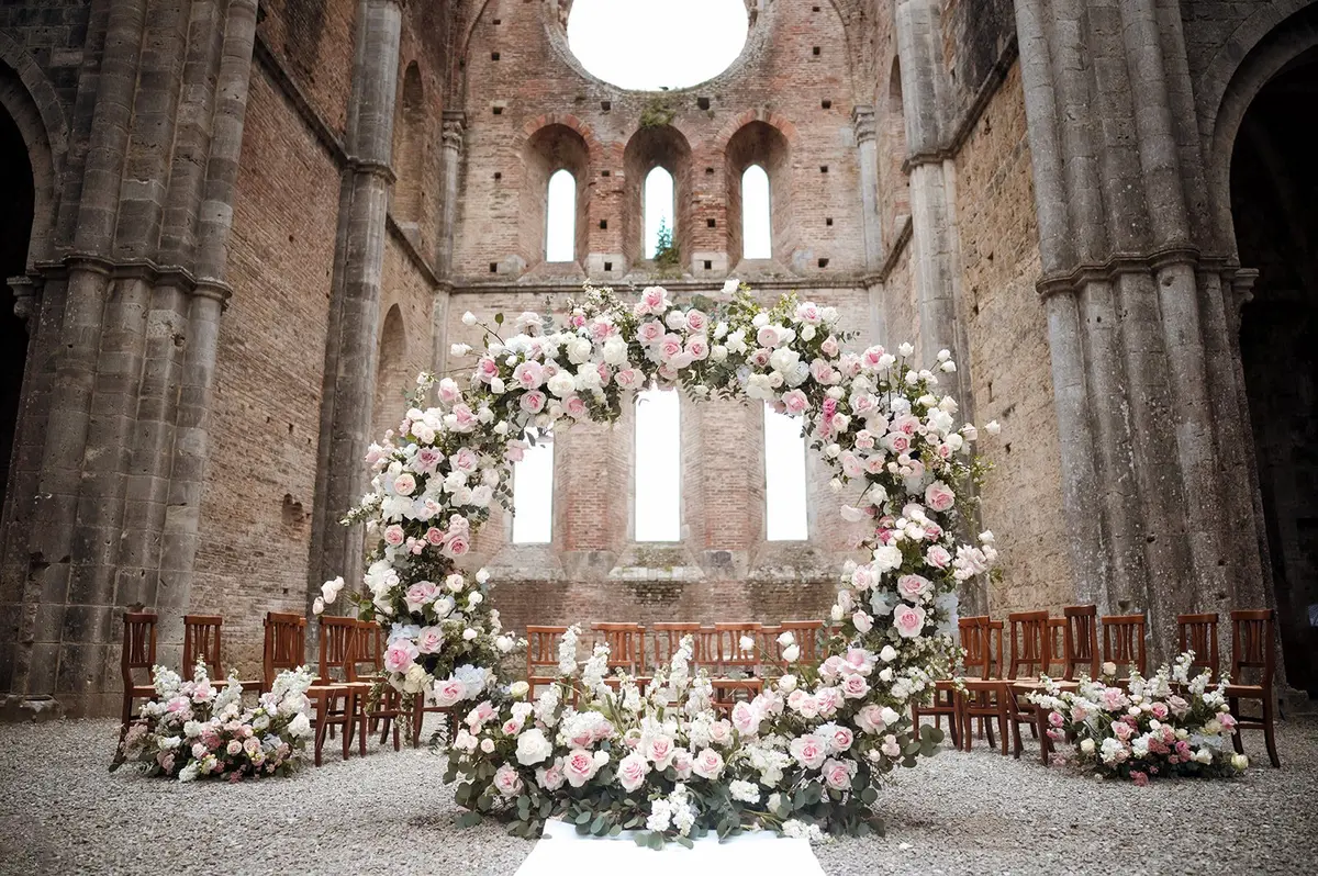 A circular arch of pink and white roses inside the roofless nave of San Galgano Abbey, Siena