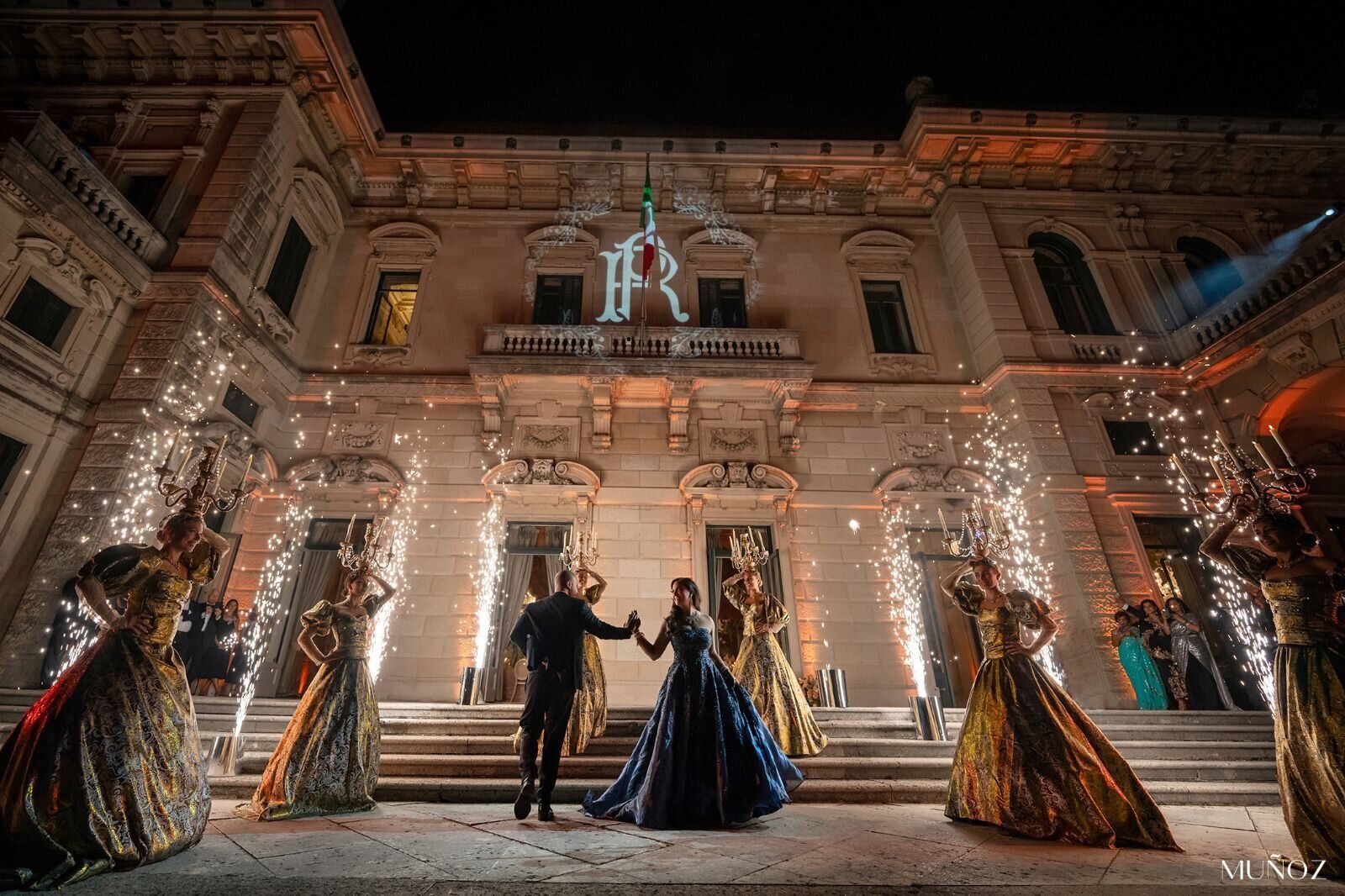 A couple dance through twin fountains of gold sparklers as gowned performers raise lit candelabra headdresses before a floodlit facade