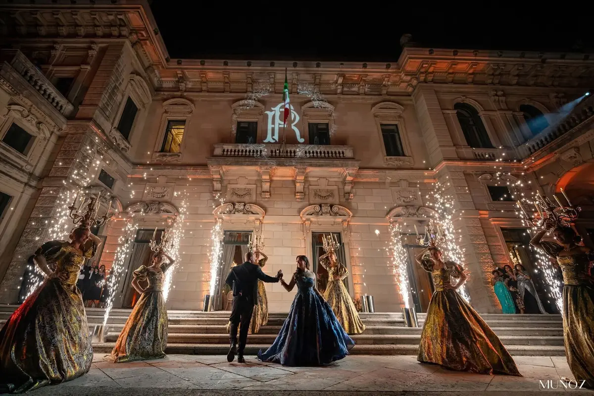 A couple dance through twin fountains of gold sparklers as gowned performers raise lit candelabra headdresses before a floodlit facade