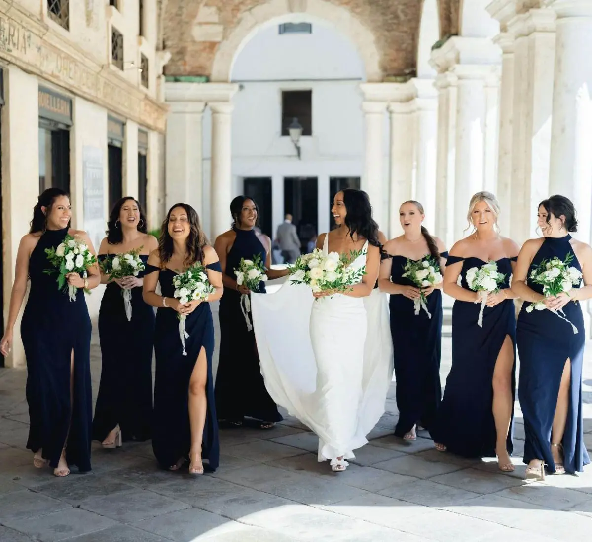 A bride and seven bridesmaids in navy gowns walking beneath a colonnaded Venetian portico