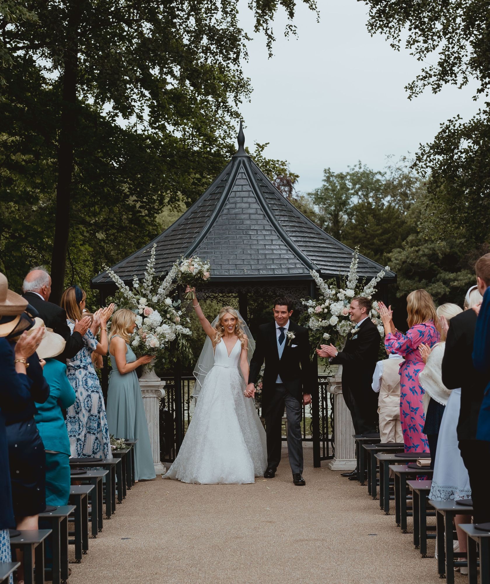 Newlyweds walking back down the outdoor aisle as guests cheer, the garden pavilion behind