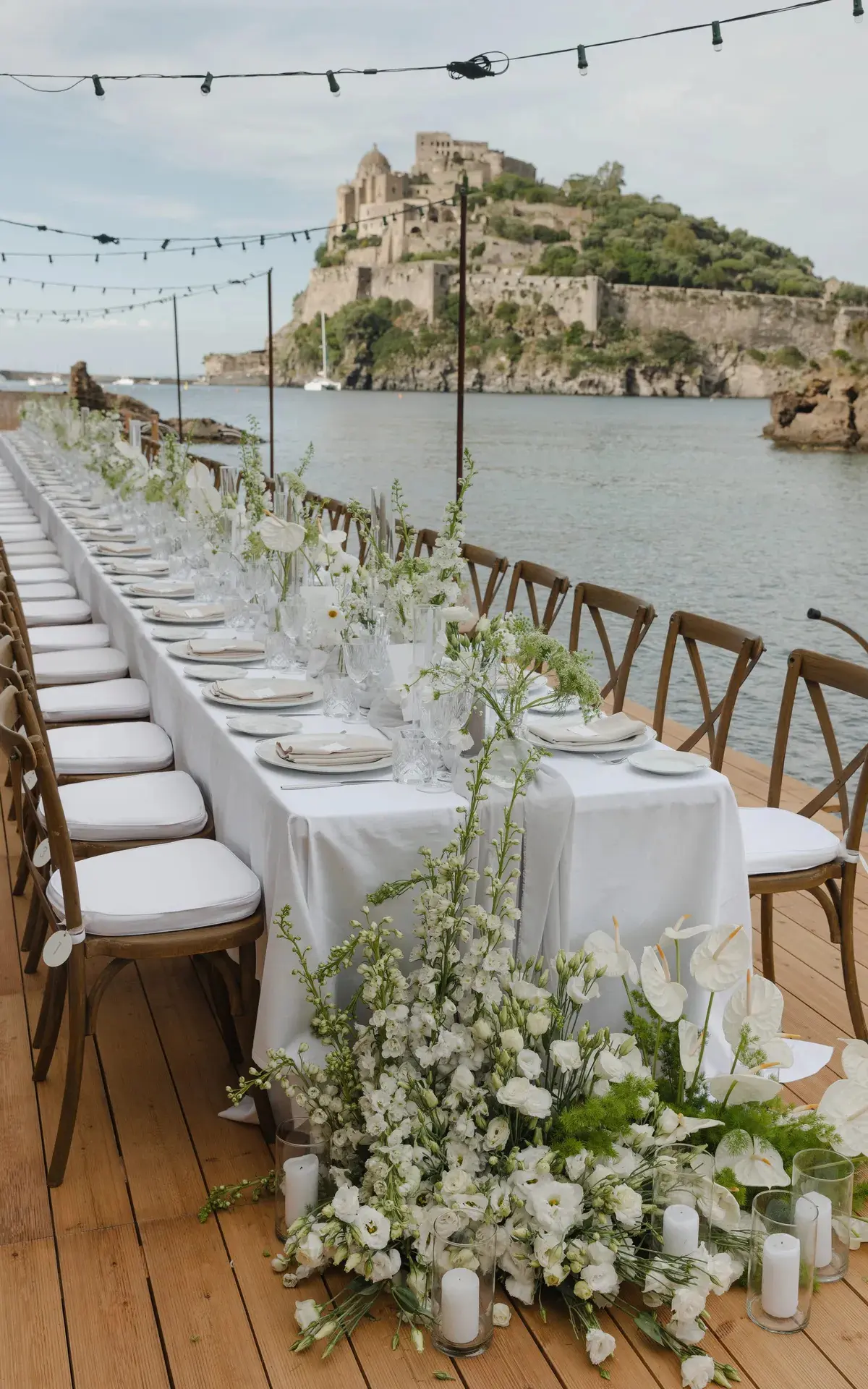 A long wedding table on a wooden deck at the water with a castle beyond