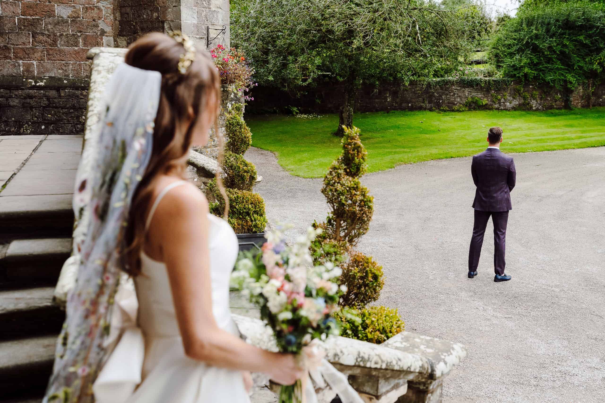 A first look in the gardens, the bride with her bouquet by clipped topiary as the groom waits
