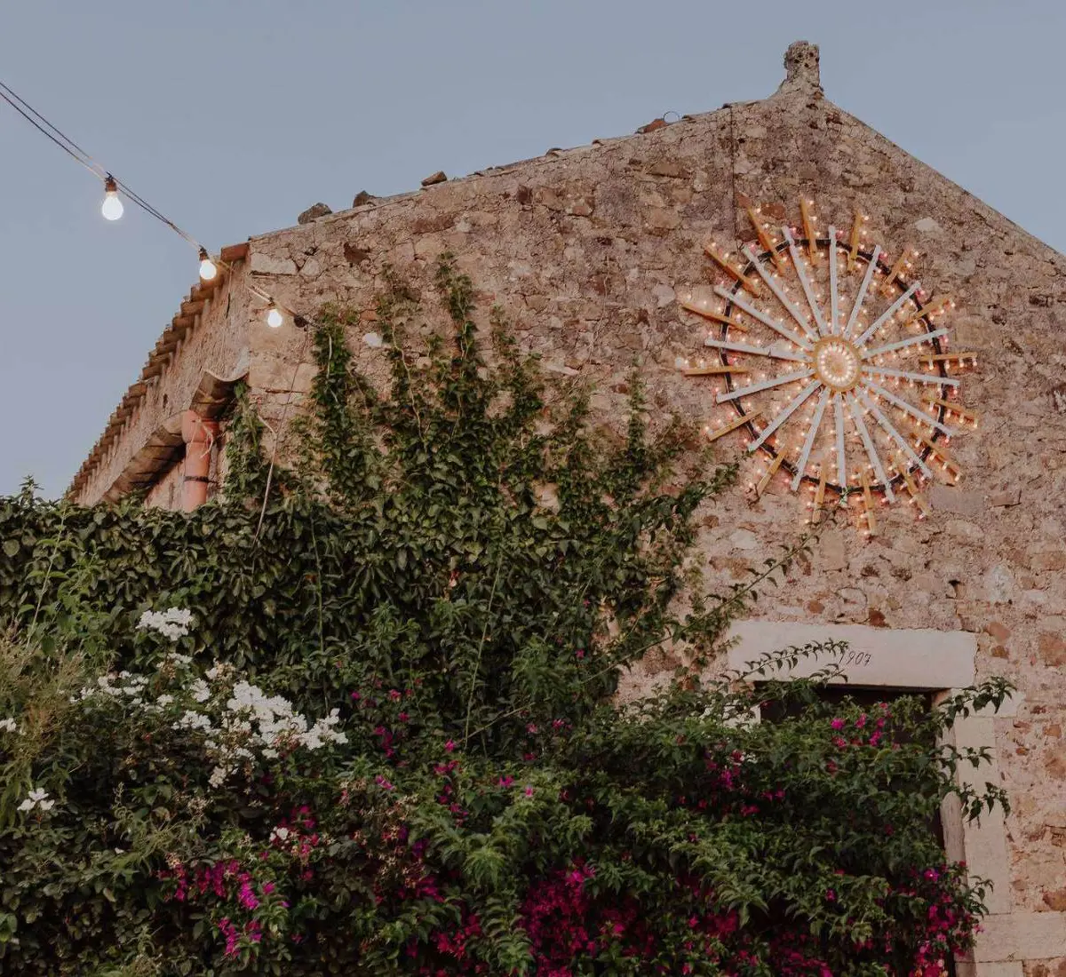 A Sicilian-style stone masseria facade at dusk, string lights and a lit sunburst wall ornament