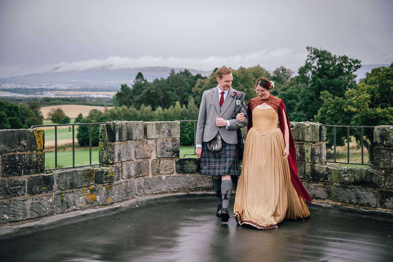 A couple, the groom in Highland dress, on a castle terrace with the hills beyond