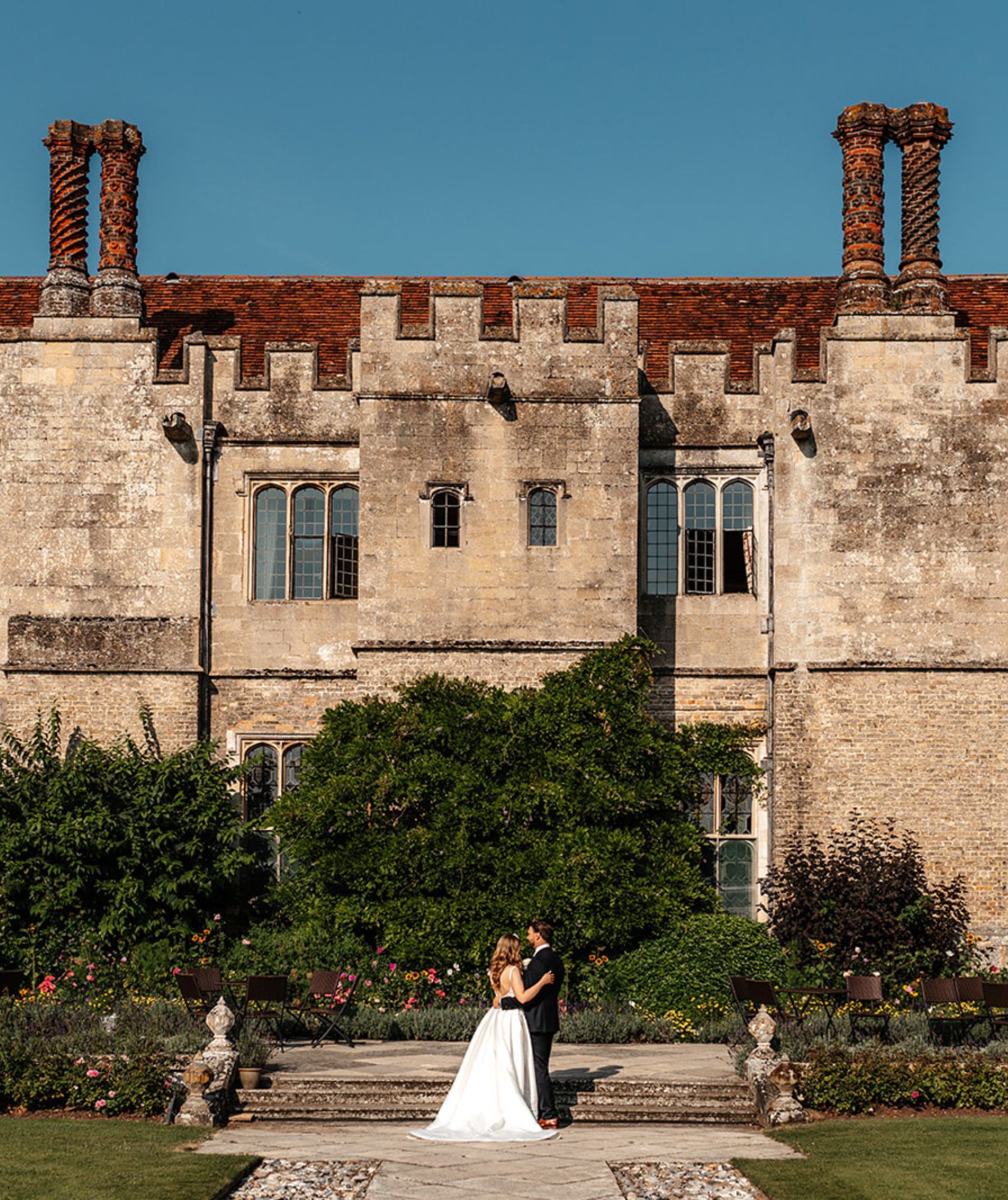 A couple embrace in the formal garden, the gabled Tudor facade and its chimneys behind
