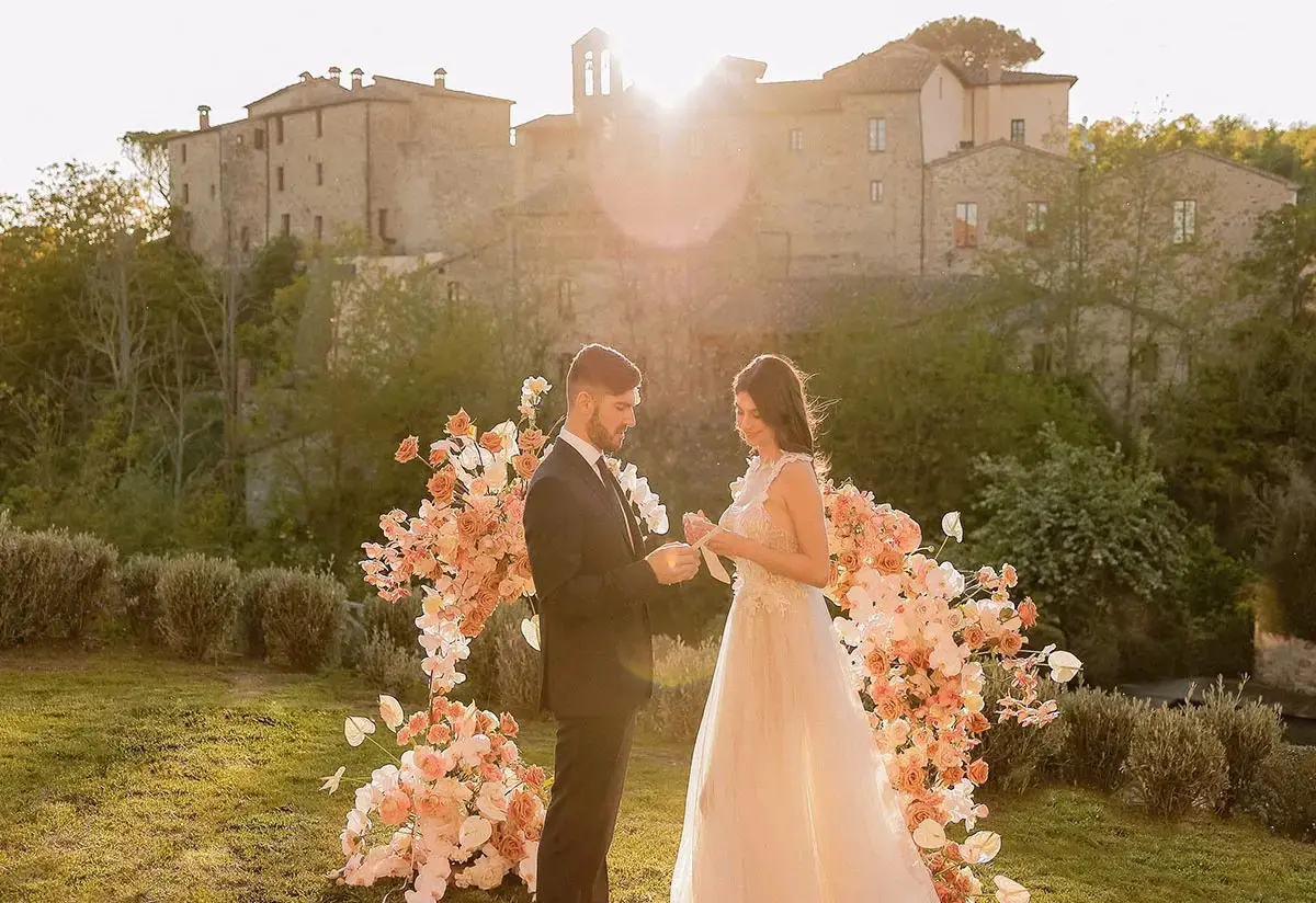 A couple exchange vows in the gardens at Castel Monastero, the castle glowing at golden hour