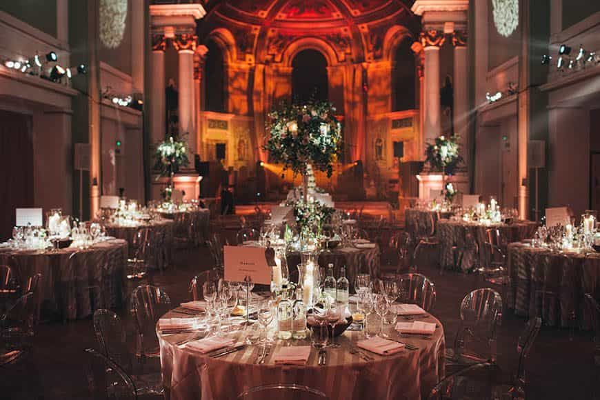 A warm amber-lit gala dinner, round tables and florals framed by the hall's soaring columns and painted apse, One Marylebone, London, England