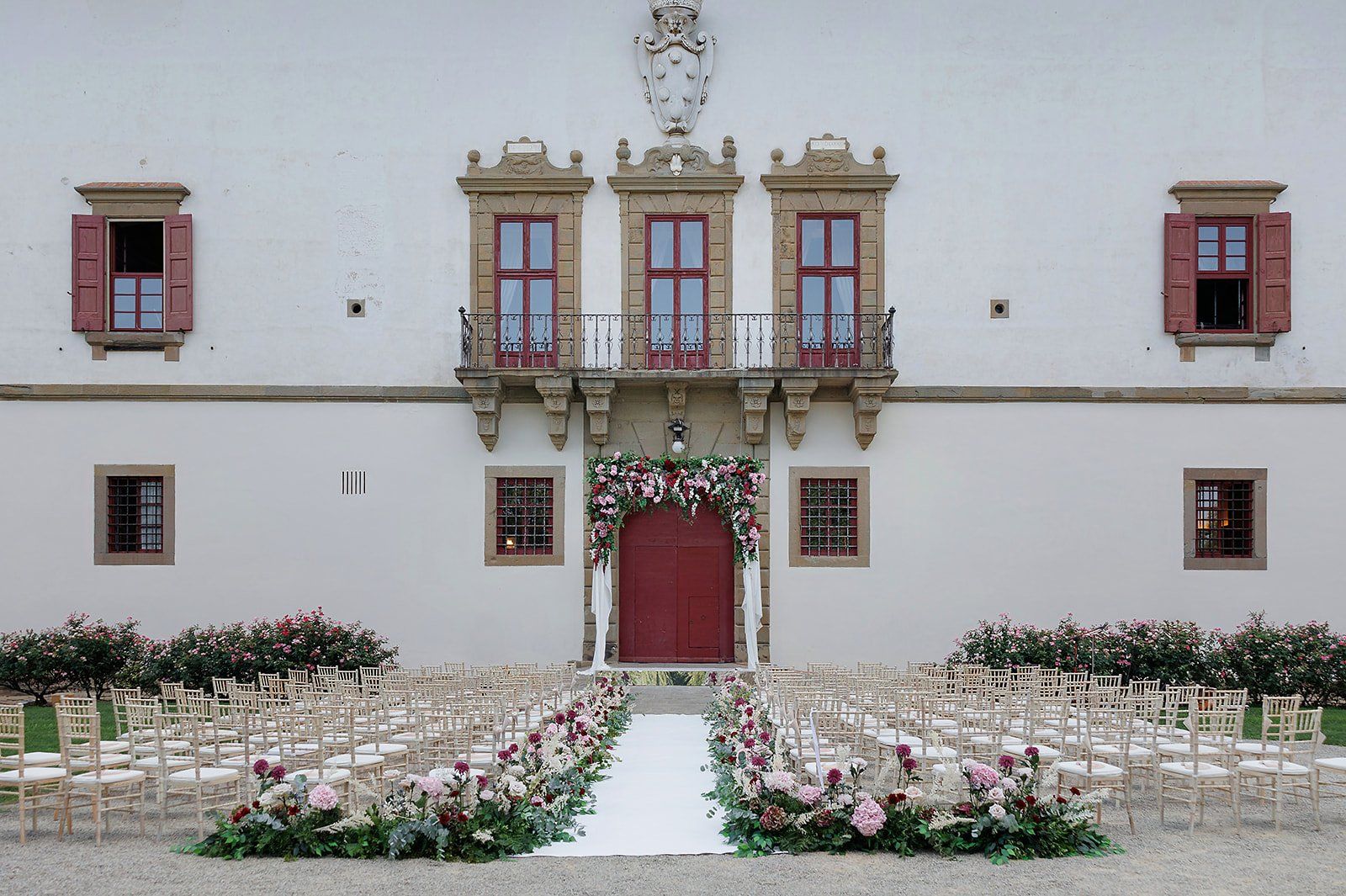The cream facade of Villa La Ferdinanda at Tenuta di Artimino, a red door under a burgundy and blush floral arch, rows of empty ceremony chairs
