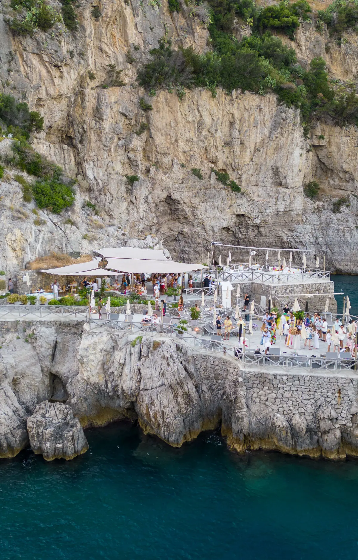 Aerial: a stone reception terrace built into the cliffs above a private cove, guests gathered under a canopy at the water's edge