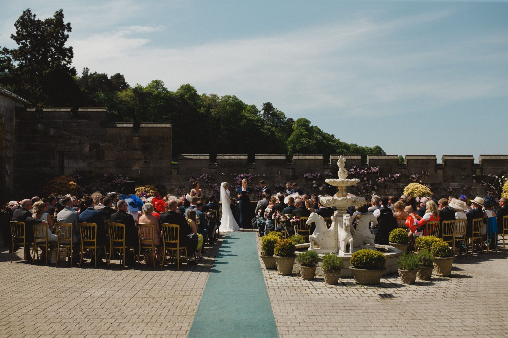 Guests seated for a courtyard ceremony around the central stone fountain, the aisle to the front