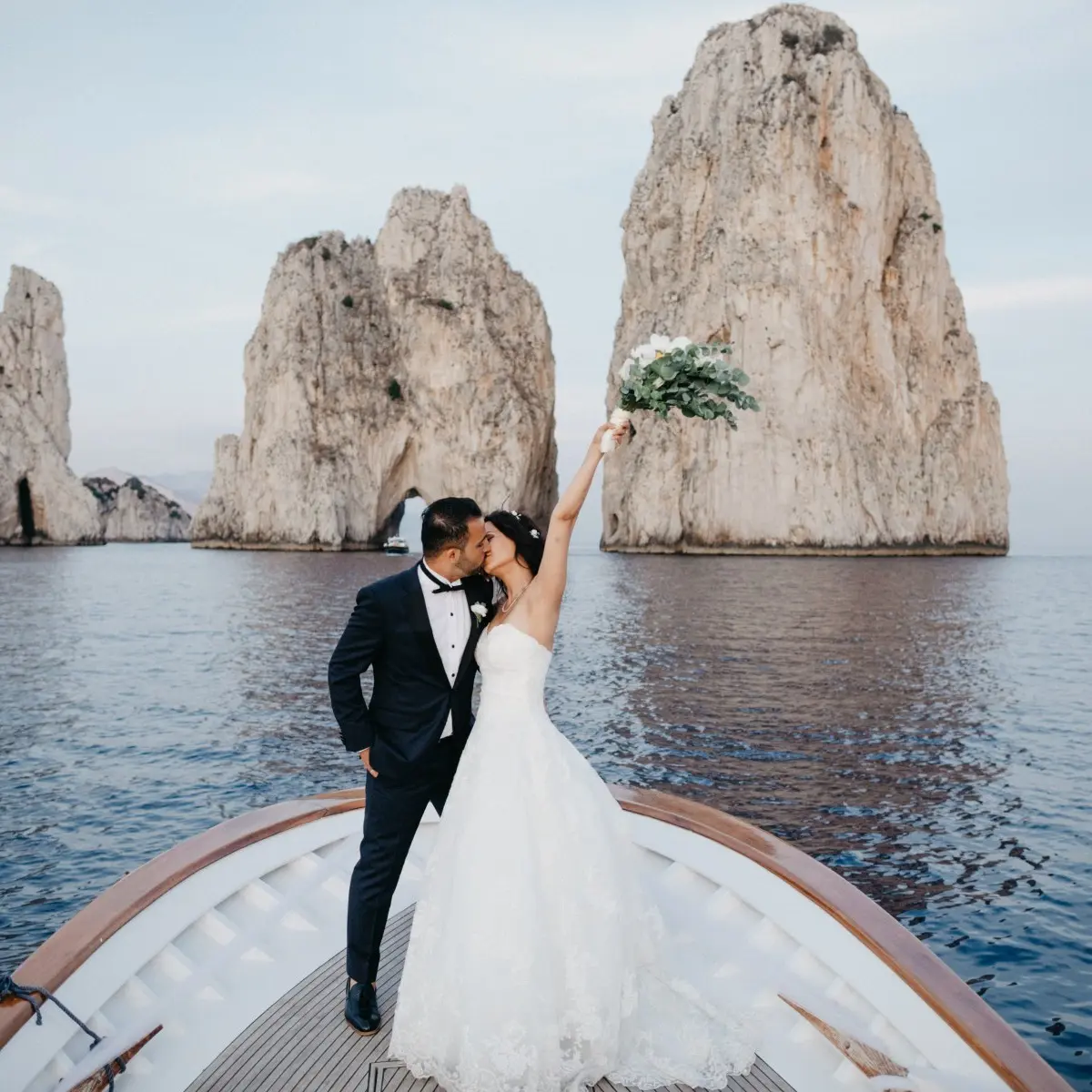 A couple kissing on the bow of a boat before Capri's Faraglioni sea stacks
