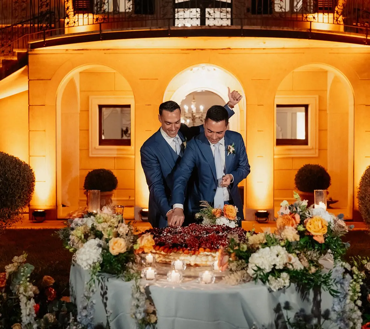 Two grooms cutting a wedding cake at night under a floodlit stone loggia
