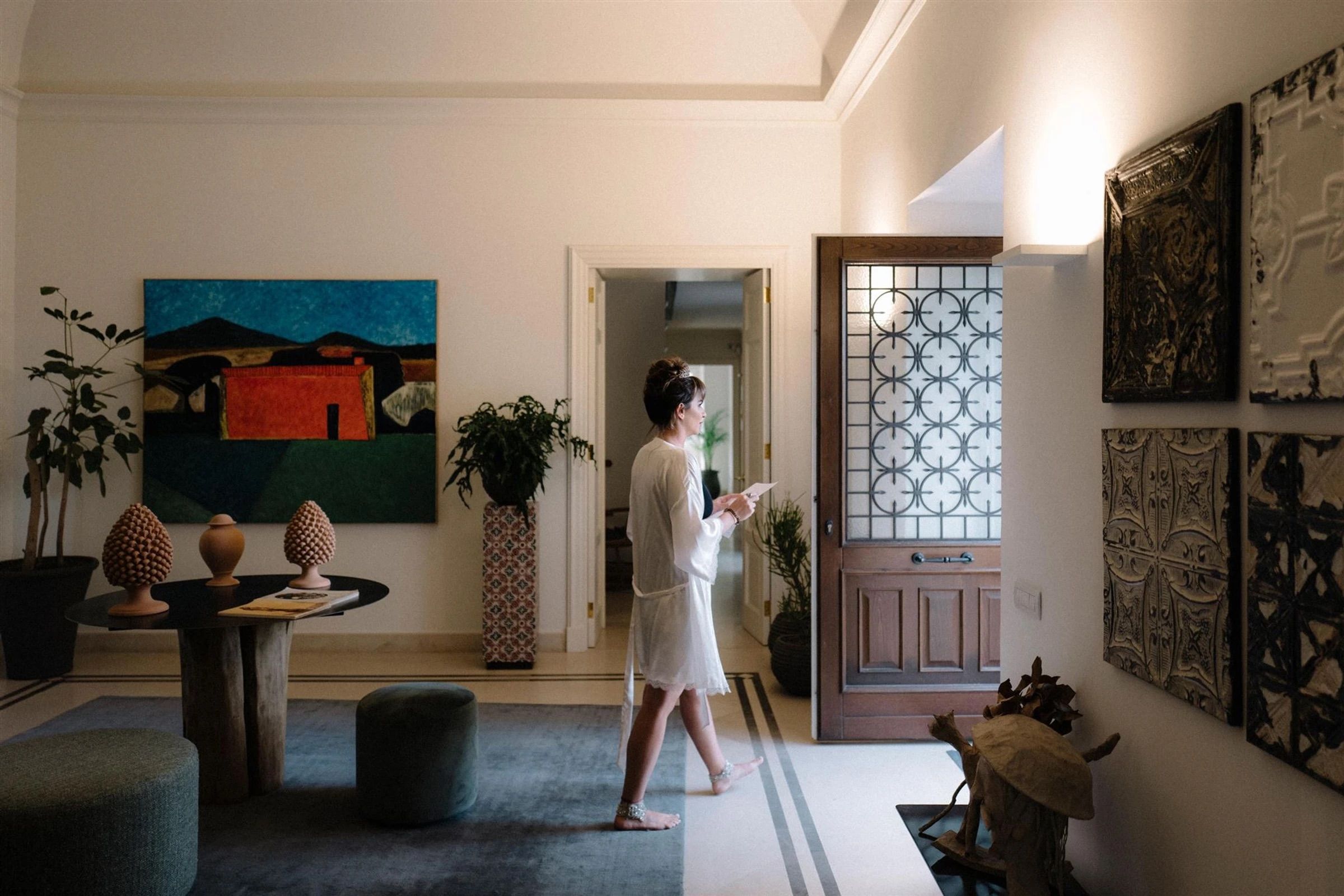 The bride, in her robe with a tiara already pinned, crosses an elegant hallway hung with Sicilian folk art