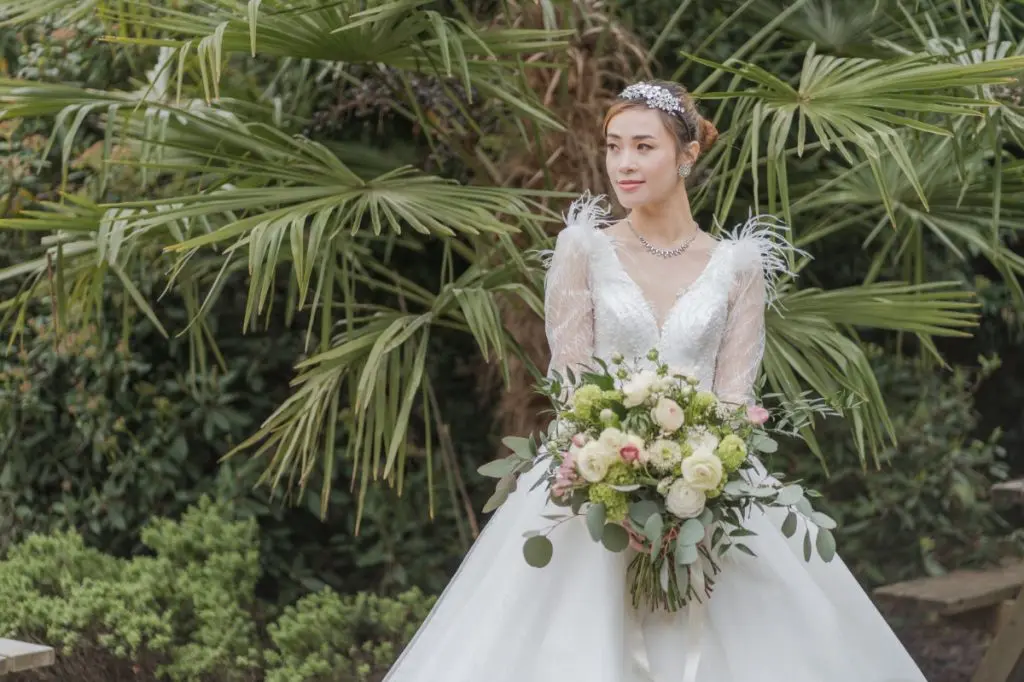 A bride in a feather-sleeve gown carries a bouquet of white rose and ranunculus, dressed with trailing eucalyptus, Shilpa Reddy, London, England