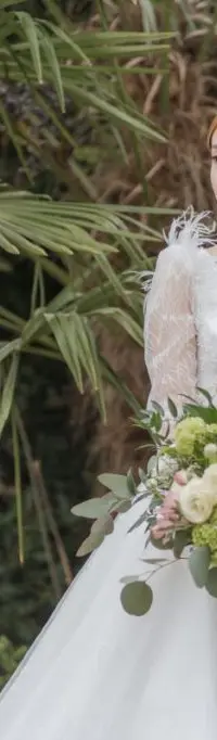 A bride in a feather-sleeve gown carries a bouquet of white rose and ranunculus, dressed with trailing eucalyptus