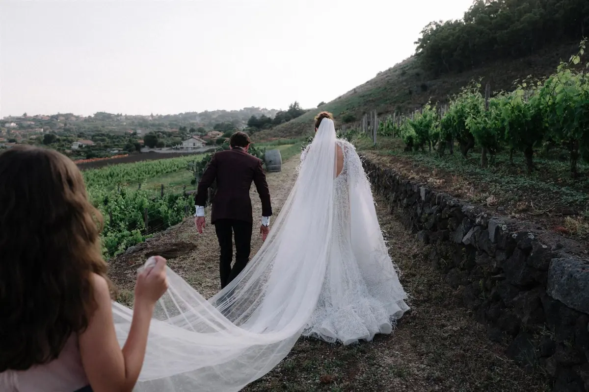 A bride and groom walk hand in hand along a dry-stone wall between rows of Etna vineyard, her cathedral veil carried behind her by a bridesmaid