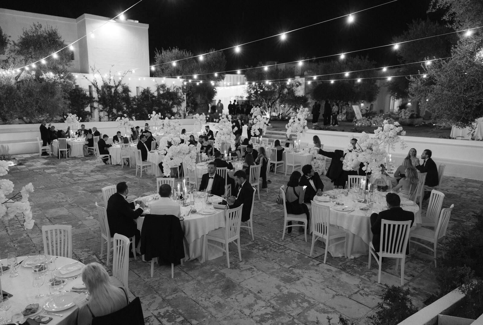 The reception in black and white, guests seated under strung lights, Puglia