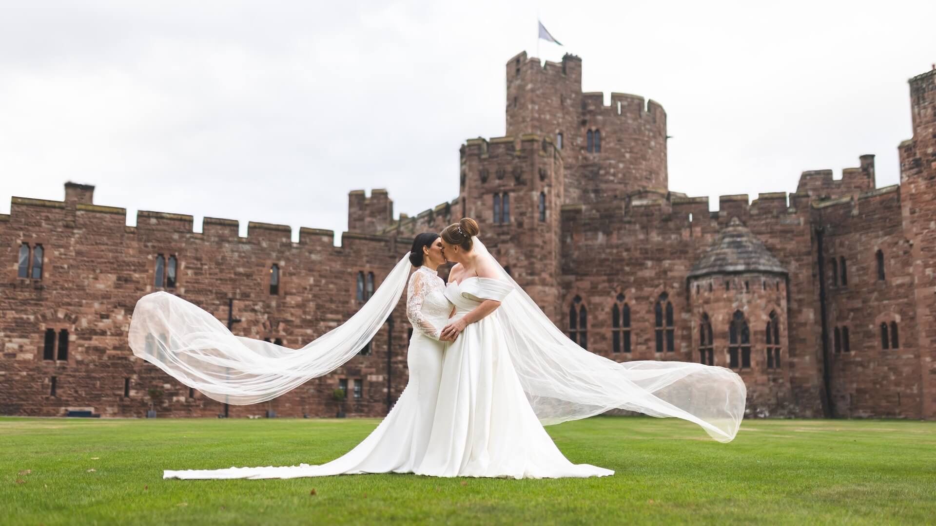 Two brides embrace on the lawn, veils sweeping, the red sandstone castle behind them