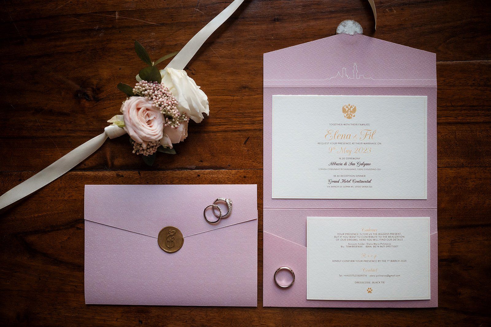 A dusty-pink wedding invitation suite naming Elena & Fil, San Galgano Abbey and Grand Hotel Continental, two rings and a blush rose boutonniere on dark wood
