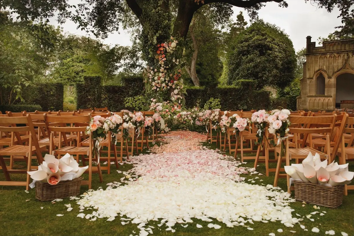 A floral arch dressed beneath an ancient tree, the ceremony aisle at Barnsley House, the Cotswolds, McQueens Flowers, London, England
