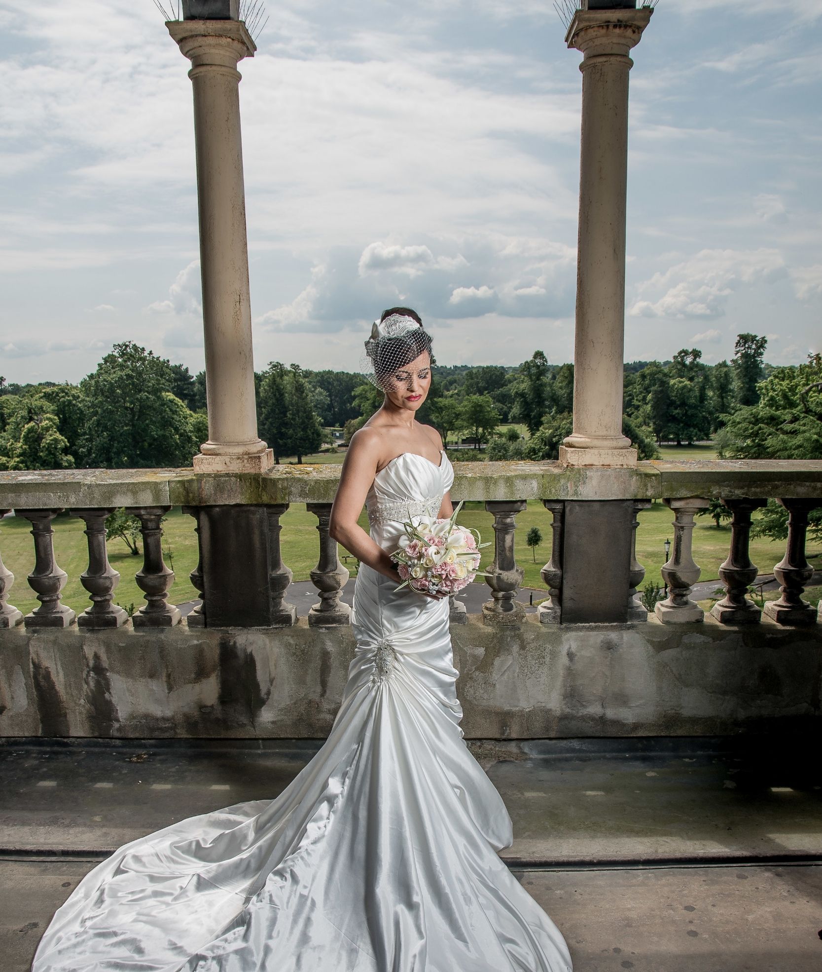 A bride in a fitted silver gown, holding her bouquet on a stone balcony