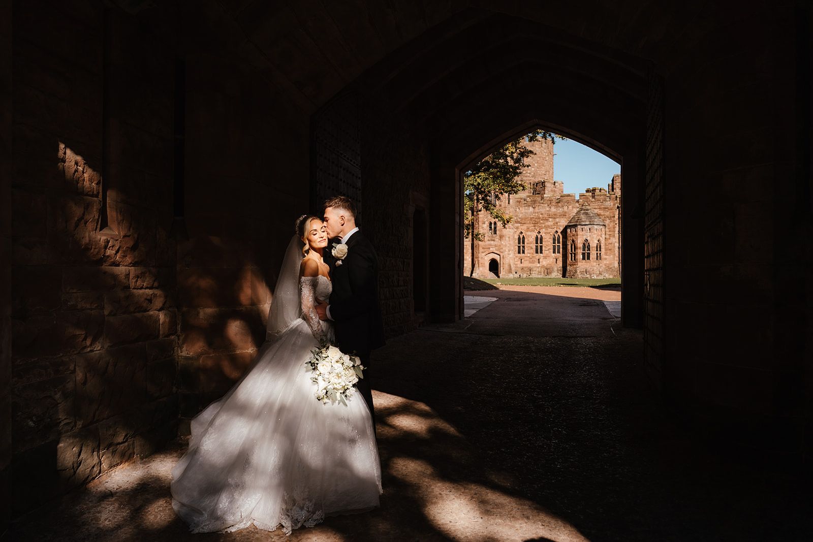 A couple in the shadowed stone gatehouse archway, the sunlit courtyard beyond