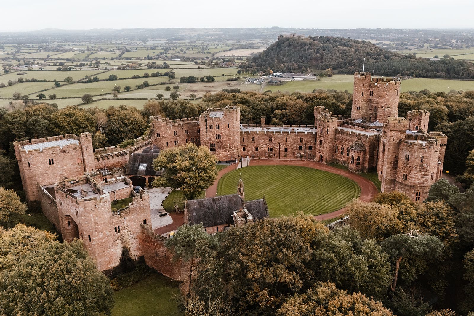 An aerial view of the whole red sandstone castle above the Cheshire plain, wrapped in autumn woodland, Peckforton Castle, Tarporley, England