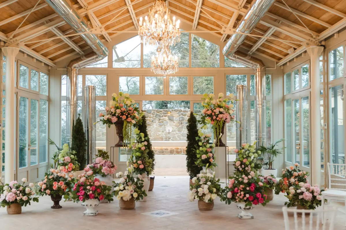 A glasshouse aisle under a timber roof, urns of pink and coral either side
