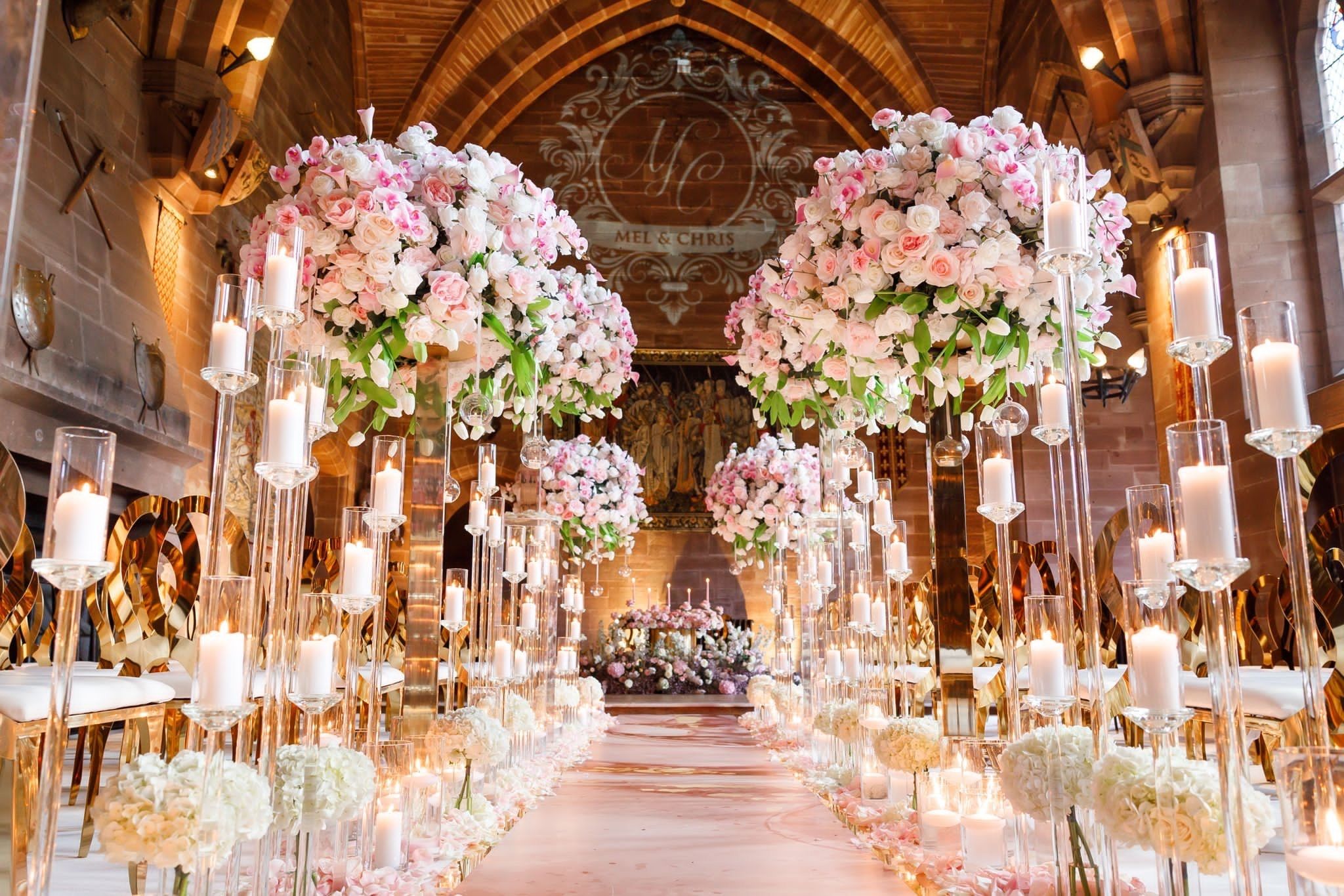 The Great Hall aisle framed by towering pink and white floral pillars and candles, a monogrammed backdrop beyond