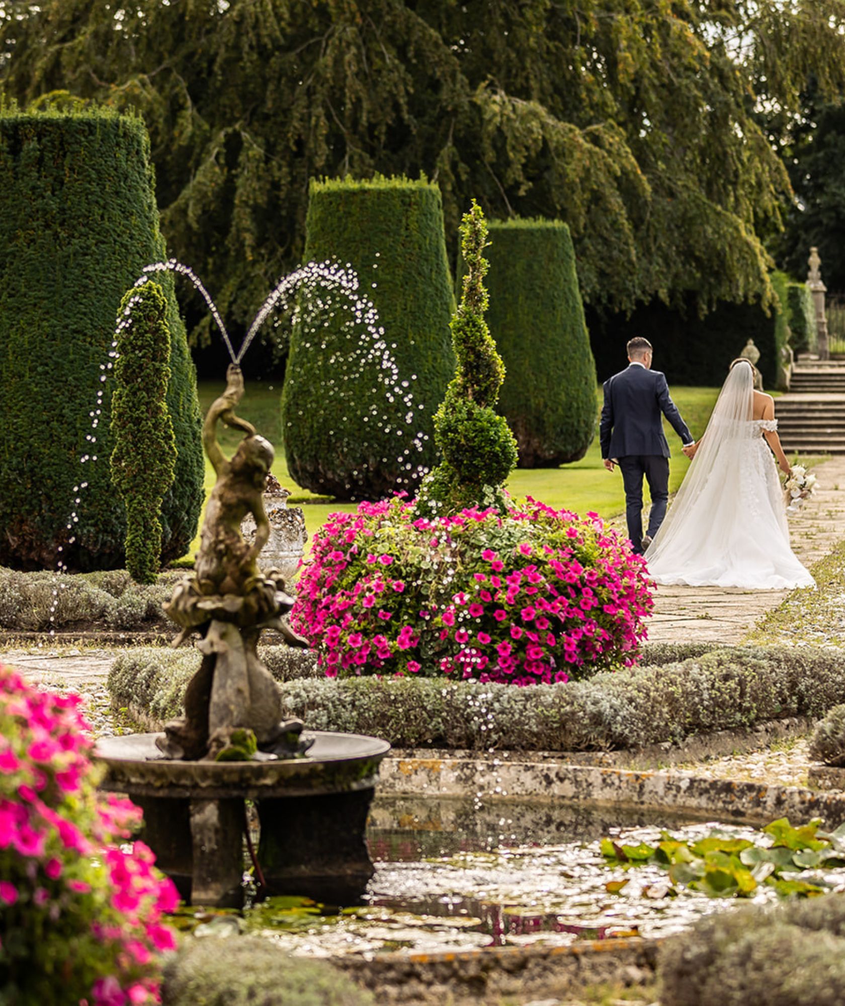 A couple walk through the topiary garden past a fountain and beds of pink flowers