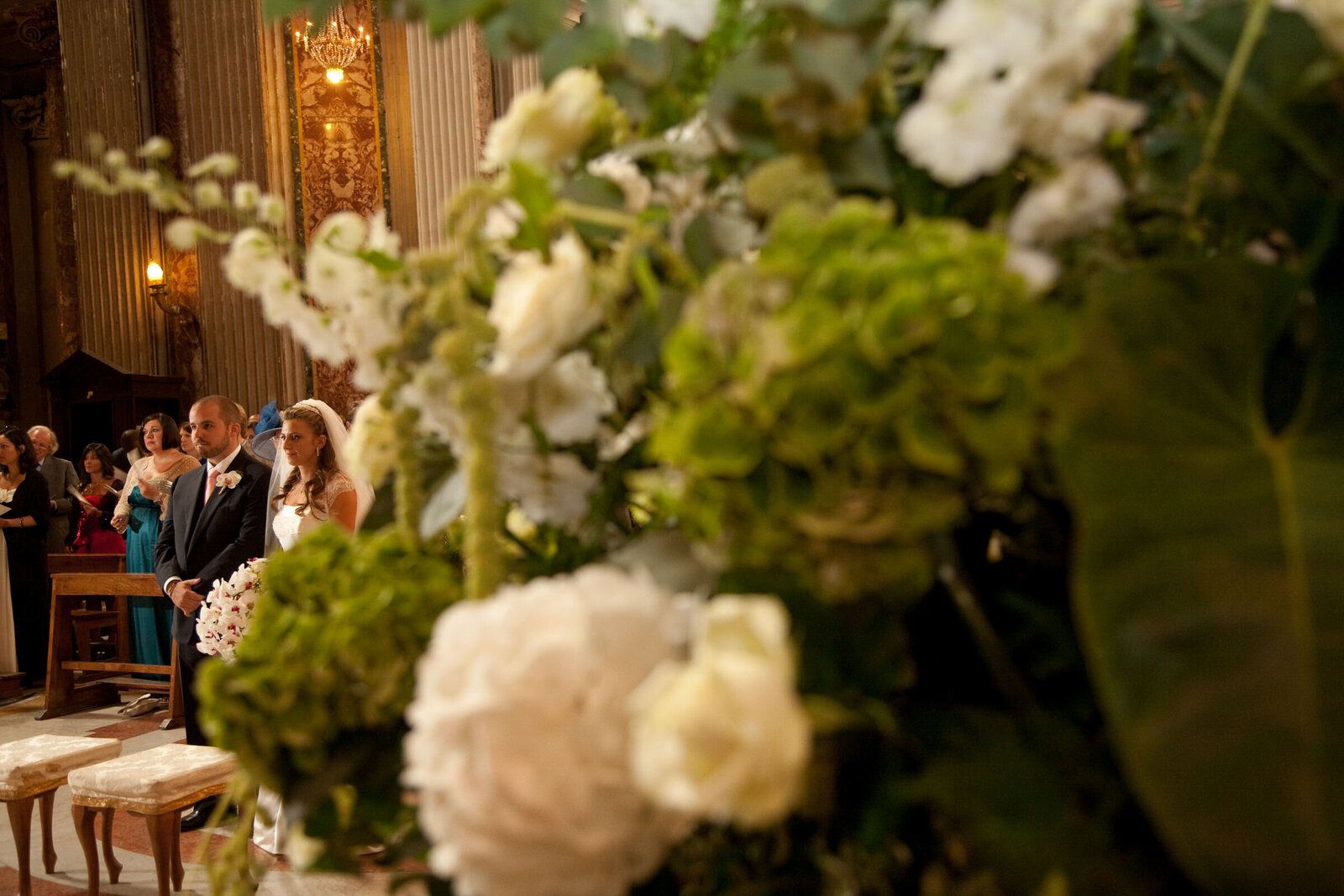 A bride and groom at the altar rail, seen through a foreground of white roses and hydrangea dressing the pew ends