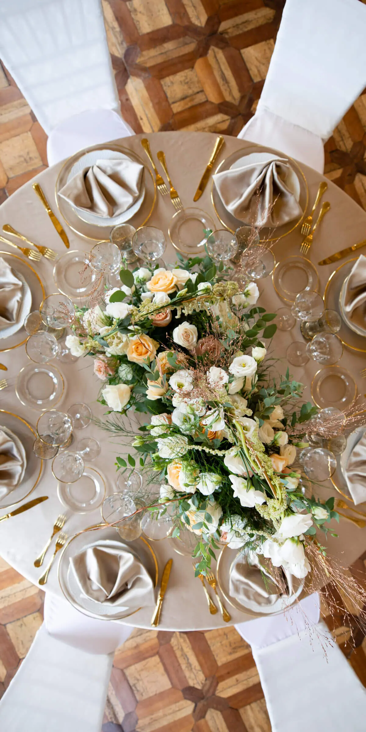 An overhead view of a round table on an inlaid parquet floor, gold flatware fanned around a low floral runner