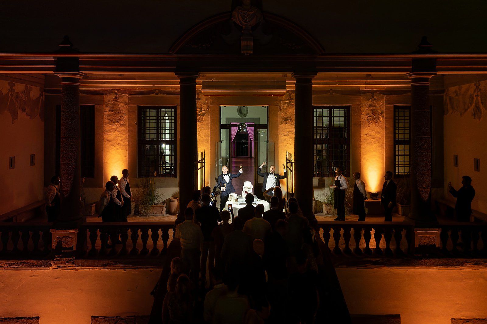 Two grooms raising champagne glasses on a candlelit, uplit loggia at night, staff and guests gathered below