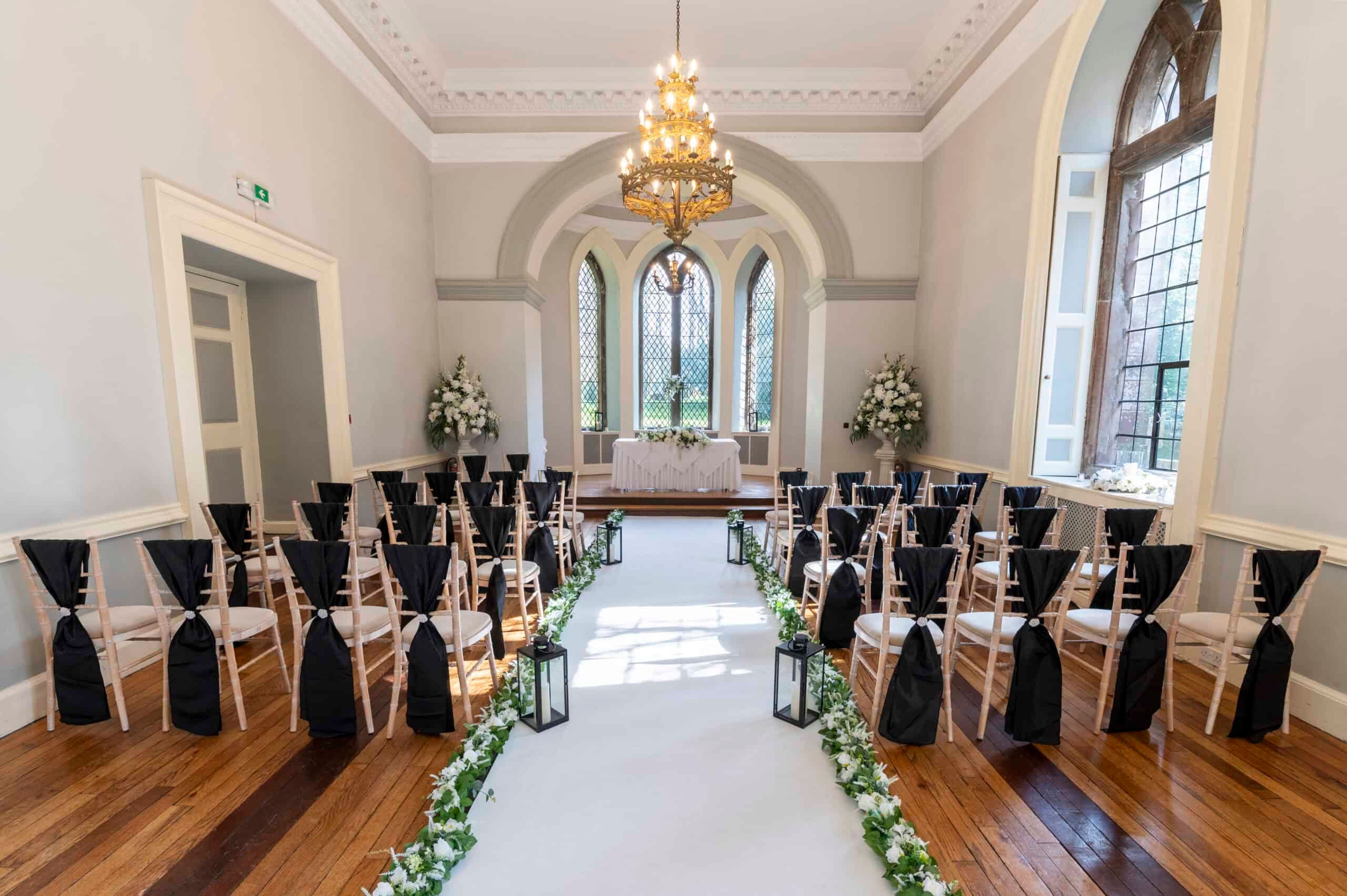 A Ballroom ceremony dressed with dark chair sashes, the aisle lit by lanterns to the arched windows