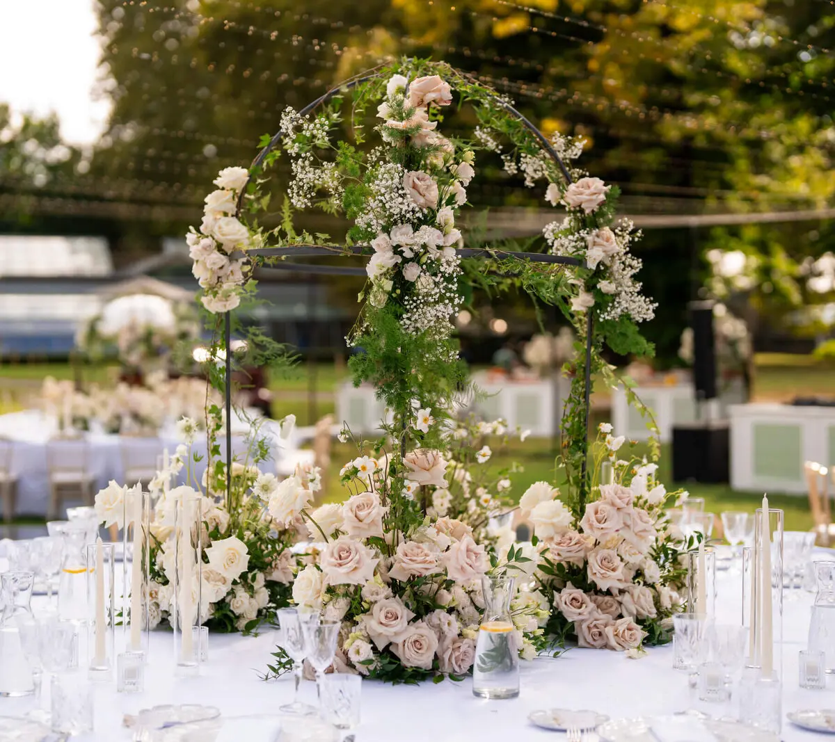 A rose-and-gypsophila arch over a linen table strung with fairy lights at dusk