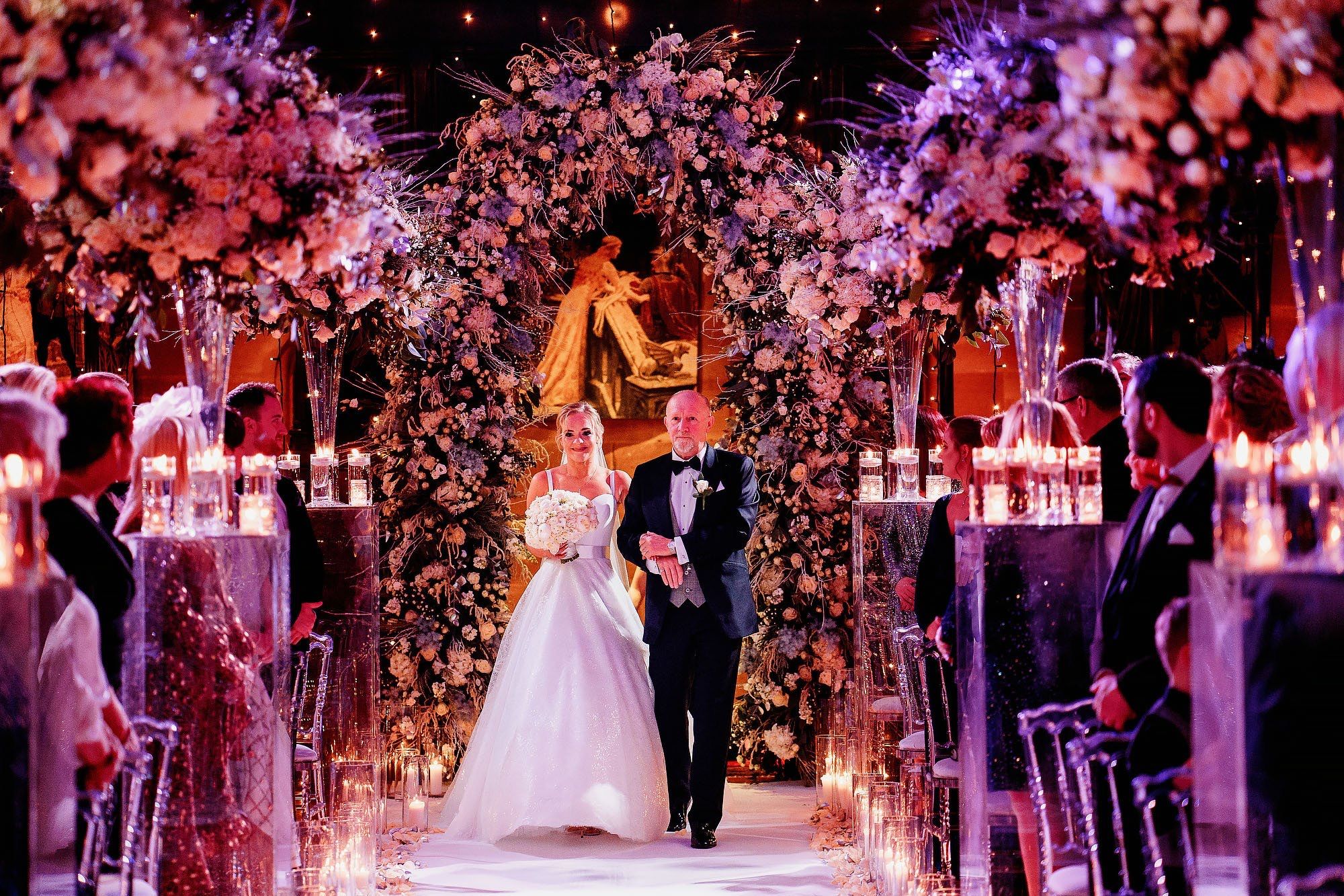A bride walks the candlelit aisle on her father's arm between blush floral arches