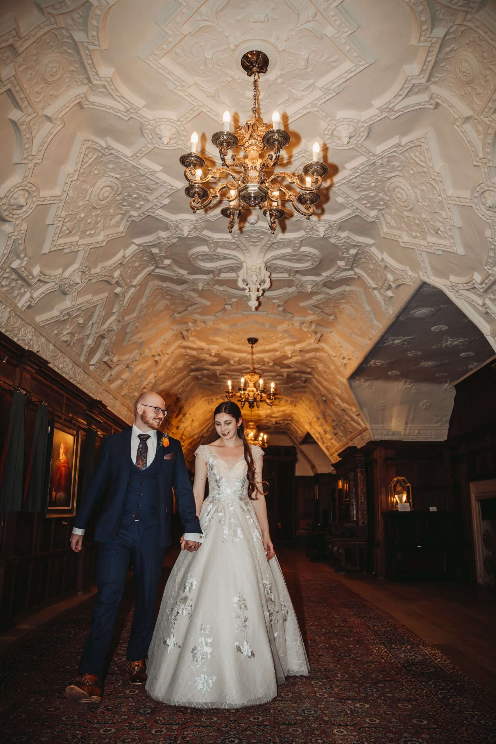 A couple beneath the ornate carved plaster ceiling and chandeliers of the castle Long Gallery